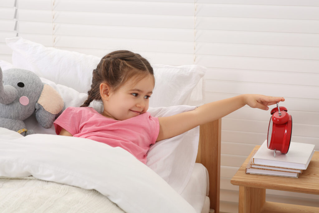 Cute little girl turning off alarm clock in cosy bedroom